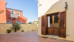 Un patio empedrado con una puerta de madera y un ventanal con contraventanas, un árbol pequeño y un edificio con balcón al fondo.