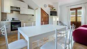Cocina compacta con armarios blancos, una mesa de comedor con sillas, un televisor sobre la encimera y un sofá rojo en la sala de estar.