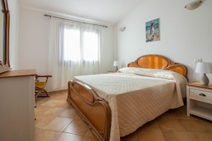 Ein Schlafzimmer mit einem hölzernen Bett, einem Nachttisch, einem Sessel, einem Schreibtisch und einem Fenster mit Vorhängen.