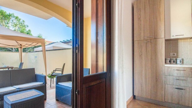 Una puerta abierta a una cocina con armarios de madera, un comedor con mobiliario de mimbre y un patio con una mesa y sillas cubiertas.
