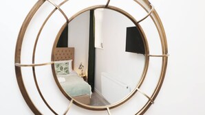 Um espelho redondo com moldura de madeira reflete um quarto com cama, mesa de cabeceira e televisão presa na parede.