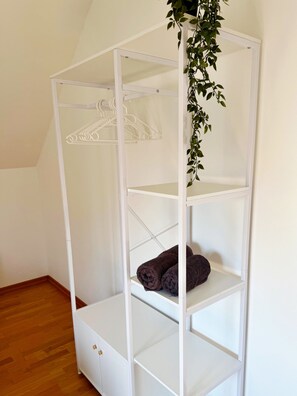 Eine weiße, mehrstufige Aufbewahrungseinheit mit Regalen und Kleiderstange, platziert in einem Raum mit Holzboden und geneigter Decke.