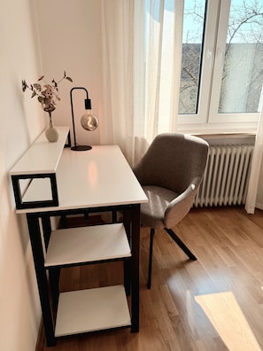 Ein modernes Zimmer mit einem weißen Schreibtisch, einem braunen Stuhl, einer Vase mit getrockneten Blumen, einer hängenden Glühbirne und einem Fenster mit Blick auf Bäume.