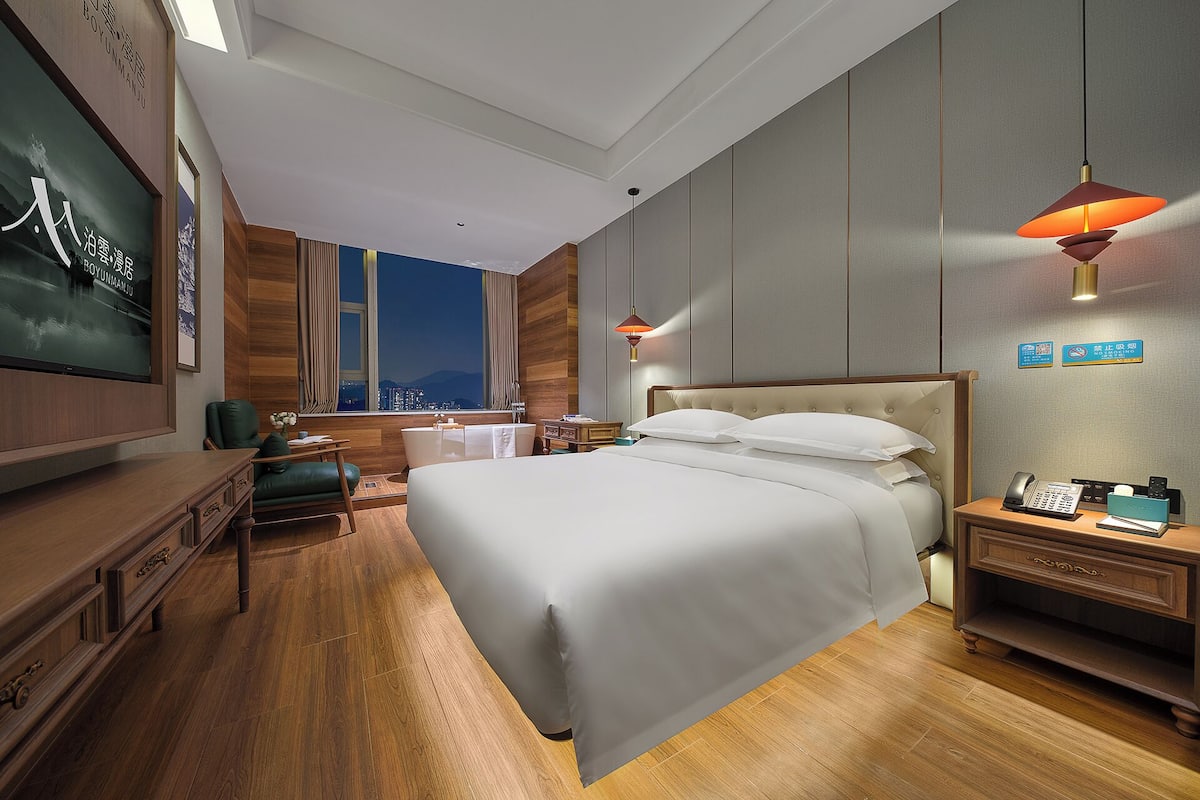 Une chambre d’hôtel avec un grand lit, un bureau, une chaise, une coiffeuse et une vue sur la ville la nuit.