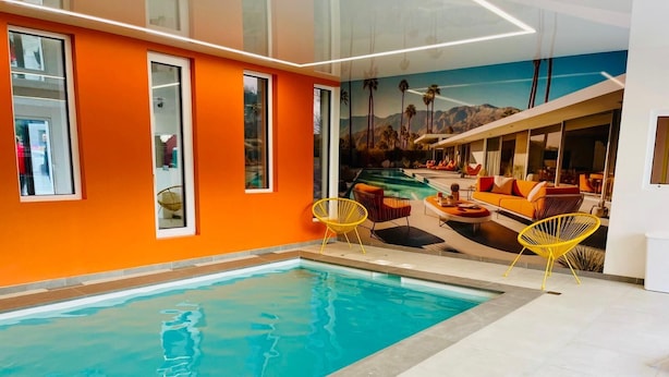 Una piscina interior con un mural de paisaje tropical, paredes anaranjadas y sillas amarillas.