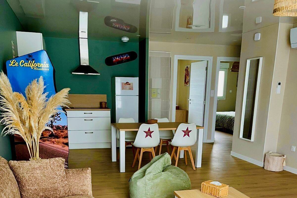 A modern living room with a sofa, a dining table with chairs, a kitchen area, and a bedroom visible through an open door.