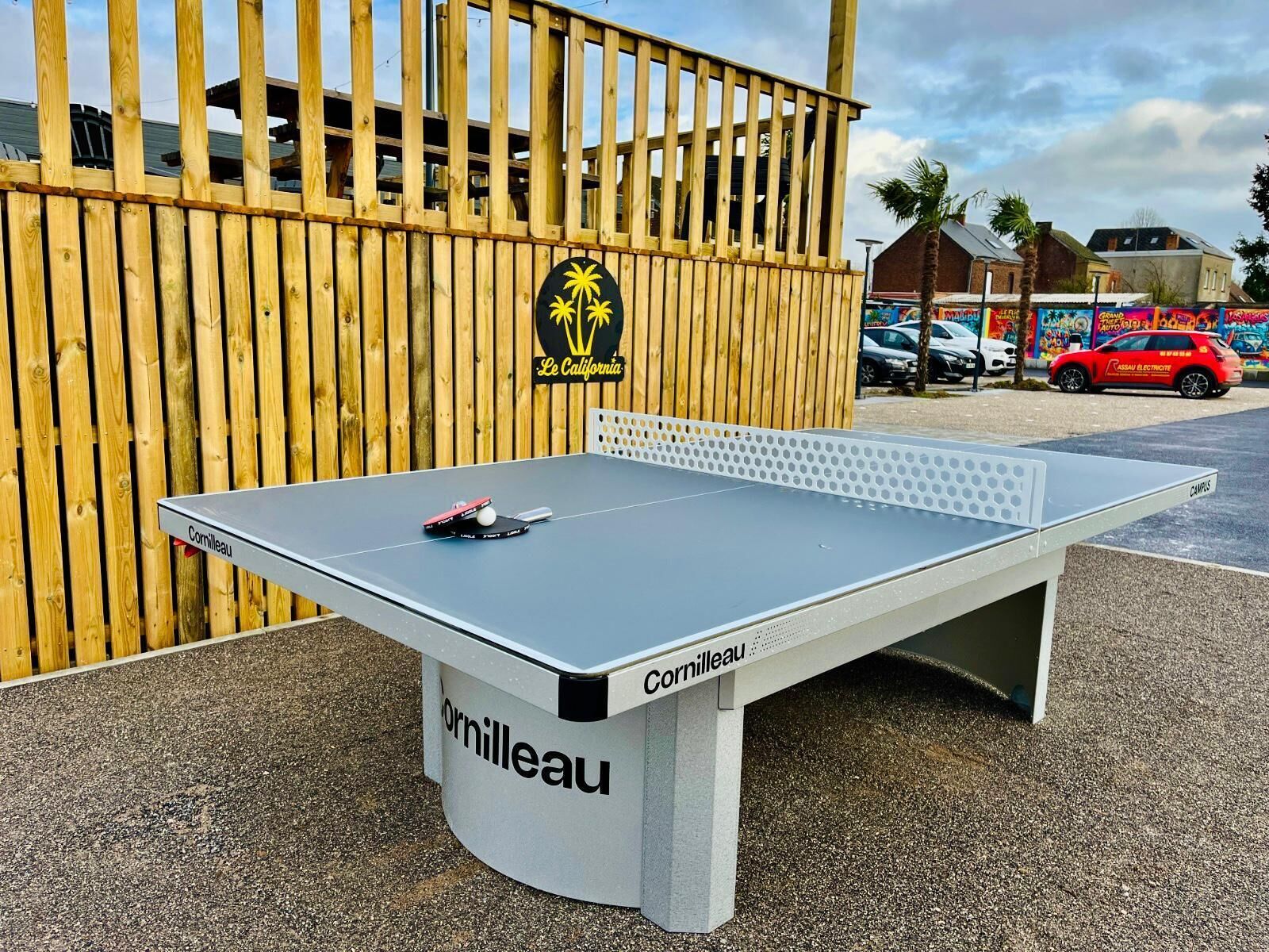 Une table de ping-pong ornée d’un logo, placée devant une clôture en bois et un bâtiment surmonté d’un palmier.
