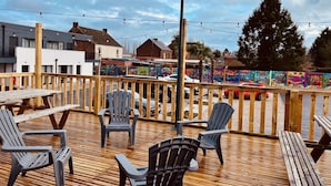 Una terraza de madera con sillas y mesas de plástico, un poste de metal y coloridas obras de arte en una cerca.