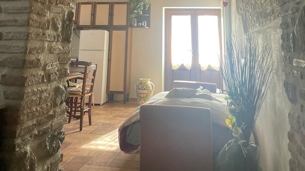 A room with a stone archway, wooden ceiling beams, a bed, a chair, a table, and a window with curtains. - Cozy apartment: Your oasis of paradise in the countryside (Anghiari)