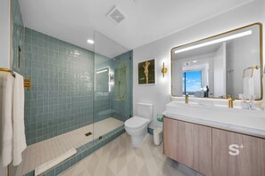 A modern bathroom with a glass-enclosed shower, a toilet, a sink with gold fixtures, and a large mirror. - 2-BD Condo in - Skyline & Ocean Views (Miami)