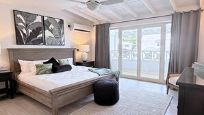 A bedroom with a bed, a ceiling fan, a nightstand, a lamp, a large window with curtains, and a view of the outside.