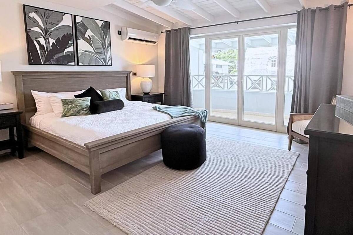 A bedroom with a bed, a ceiling fan, a nightstand, a lamp, a large window with curtains, and a view of the outside.