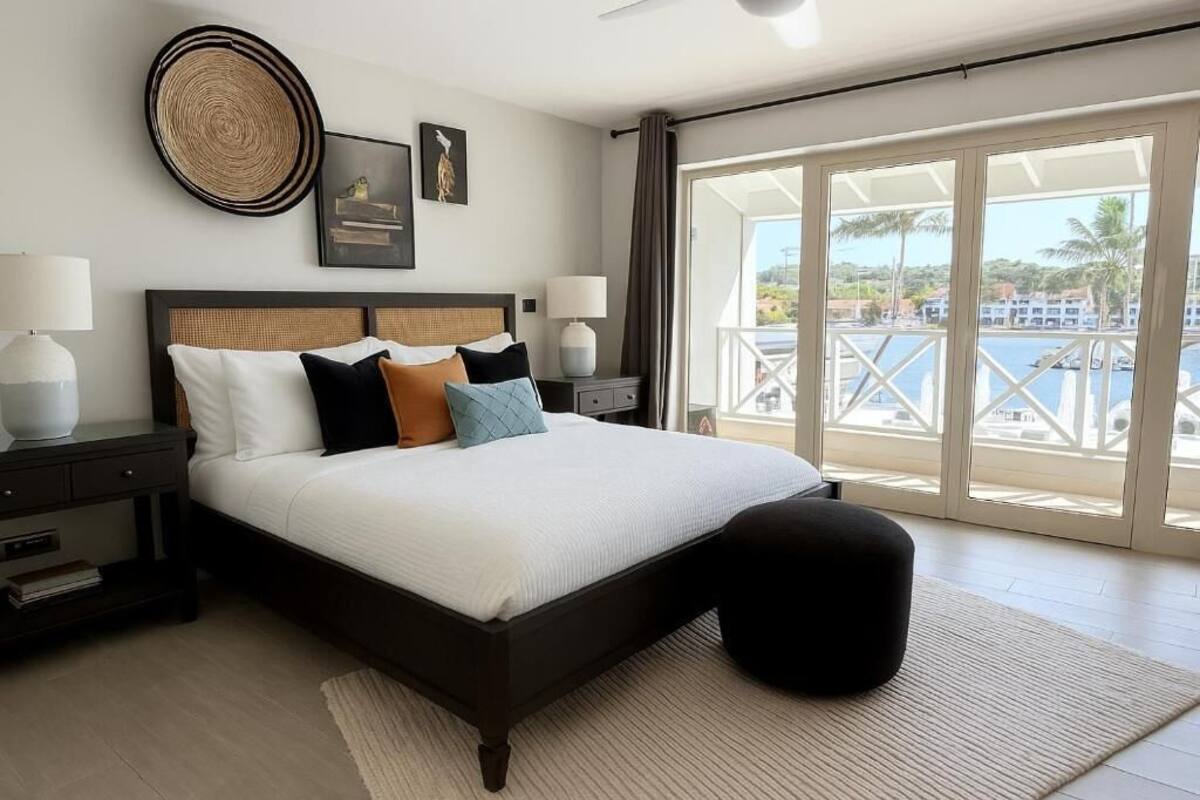 A bedroom with a large bed, a nightstand, a wall-mounted artwork, and a view of a balcony with a railing.