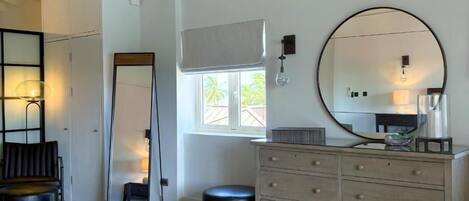 Et moderne soveværelse med et stort spejl, en kommode, et rundt spejl, et vindue med udsigt, en stol og en lampe.