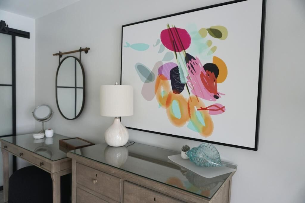 A bathroom with a glass vanity, a white vase, a framed abstract painting, a round mirror, and a towel rack.
