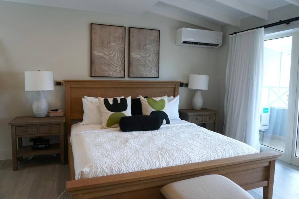 A neatly made bed with a white comforter and a wooden headboard, flanked by two bedside tables with lamps, a wall-mounted air conditioner, and a large window with curtains.