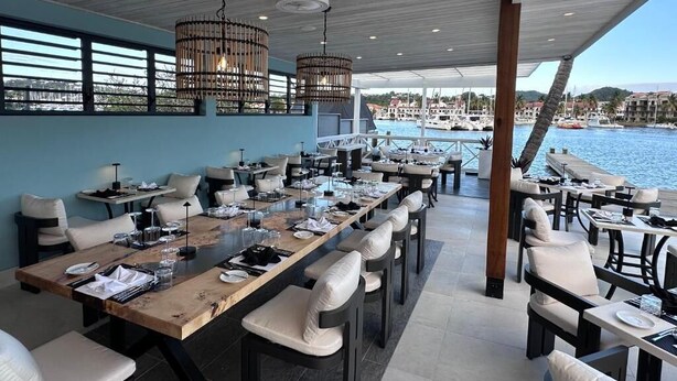A restaurant with a long table, chairs, hanging lights, and a view of the water.