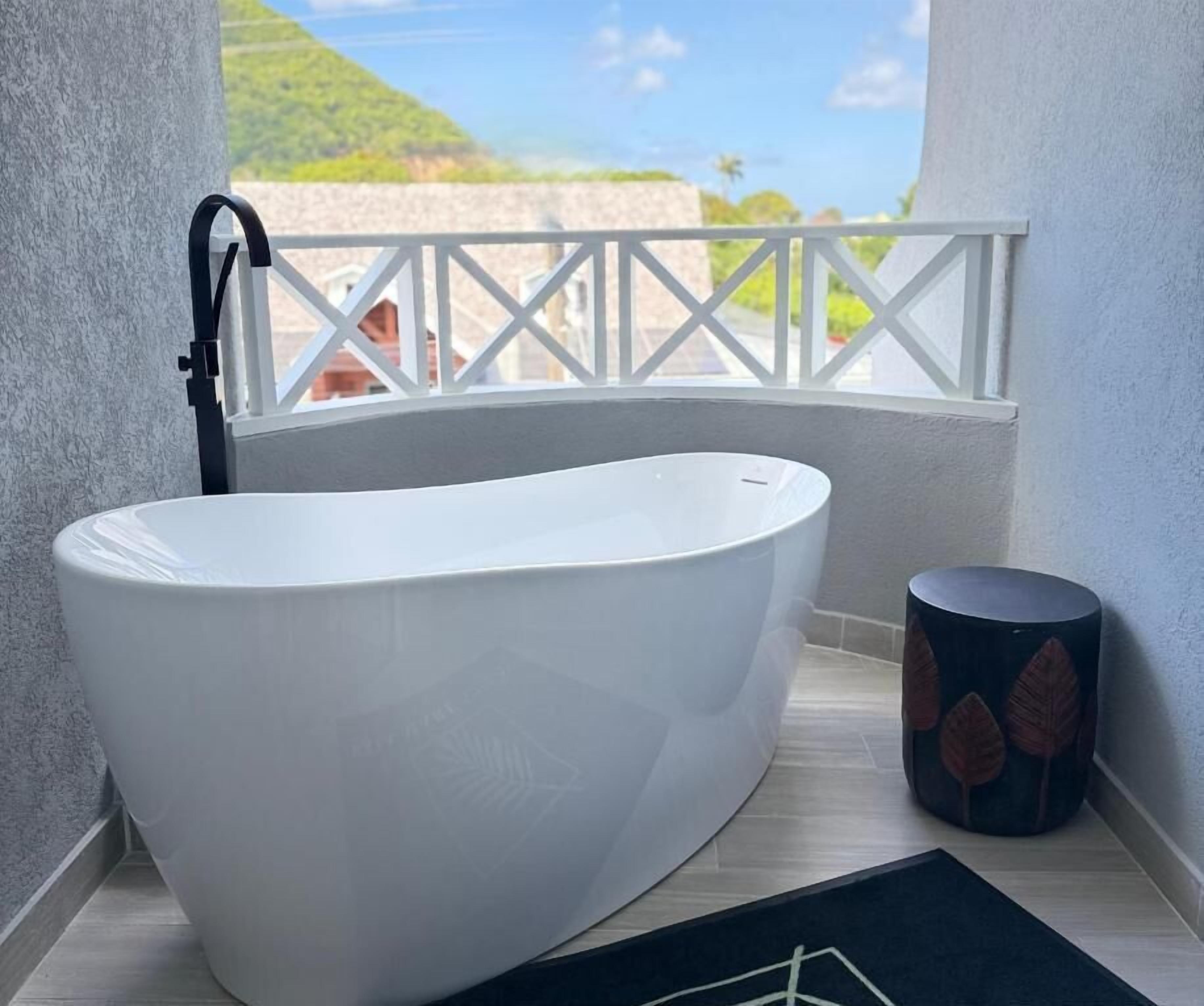 A white freestanding bathtub in a balcony bathroom with a view of a beach and a white railing.