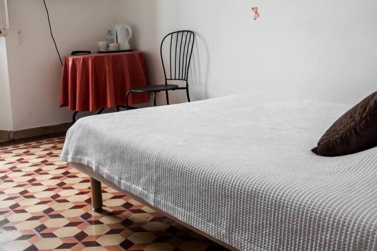 Une chambre avec un lit, un téléviseur fixé au mur, une petite table recouverte d’une nappe rouge, une chaise et des autocollants représentant des papillons sur le mur.