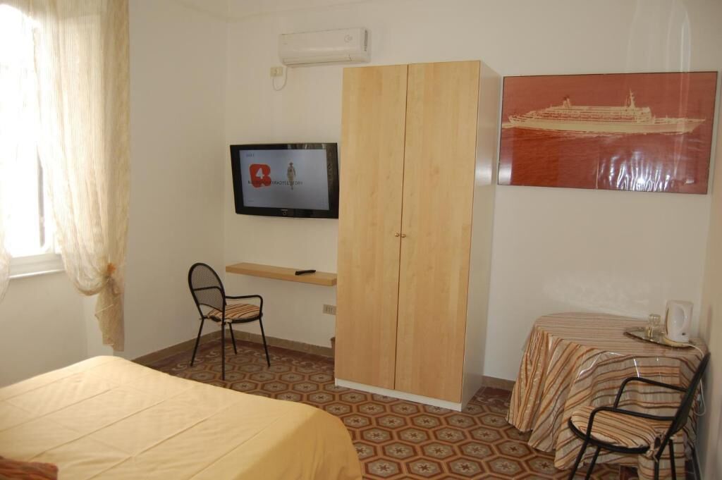 A room with a bed, a desk with a chair, a wardrobe, a television, and a table with a chair.