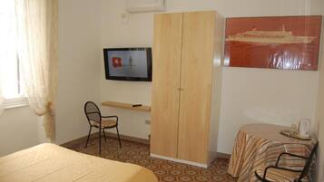 A room with a bed, a desk with a chair, a wardrobe, a television, and a table with a chair.