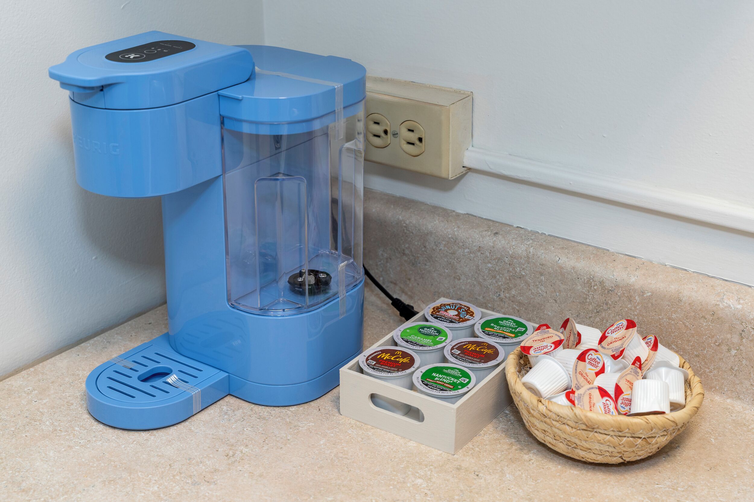 Een blauwe koffieapparaat met een transparante watertank, een mand met koffiekapsules, en een stopcontact op een aanrecht.