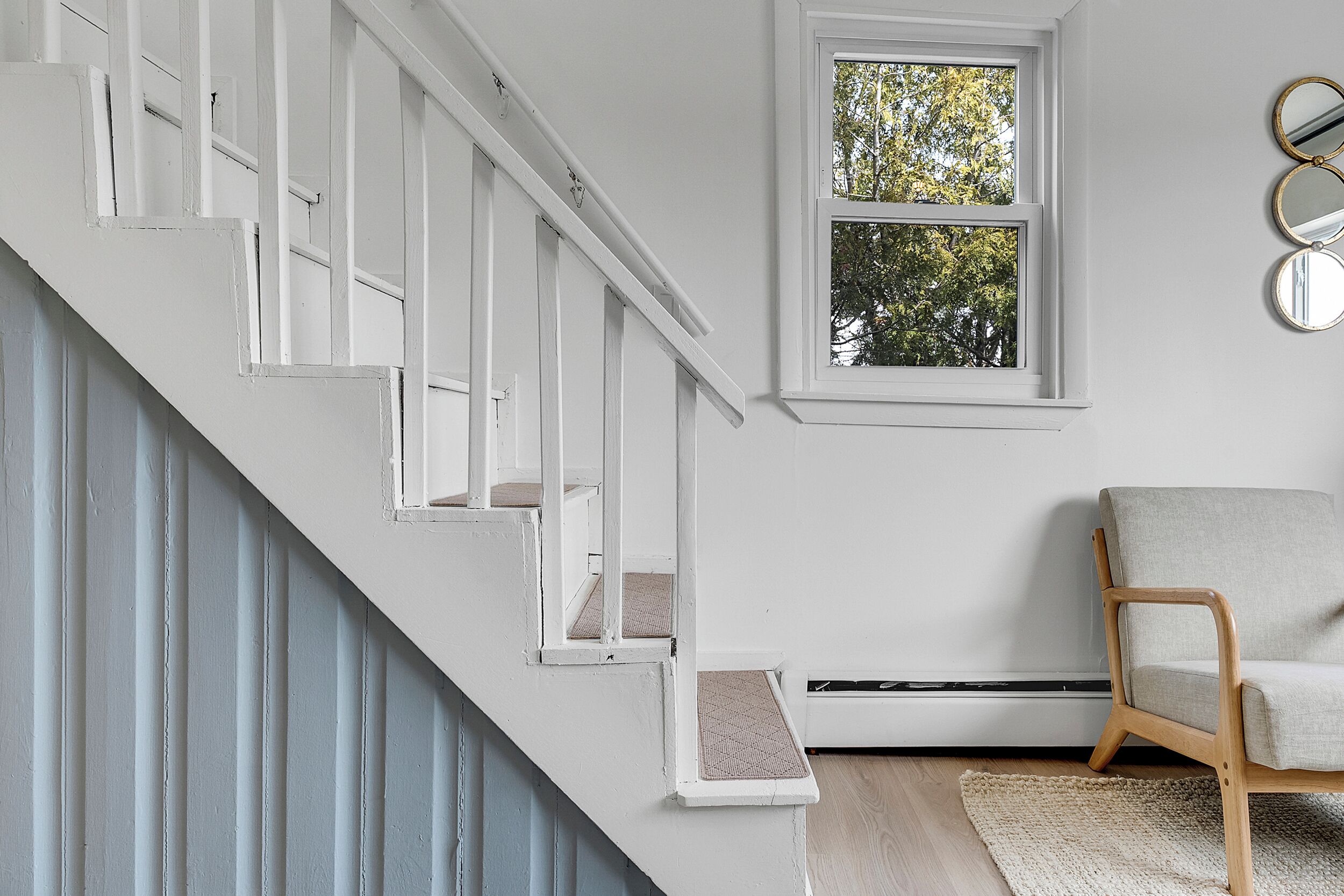 A staircase with white railings, a window with a view of trees, a chair, and a radiator.