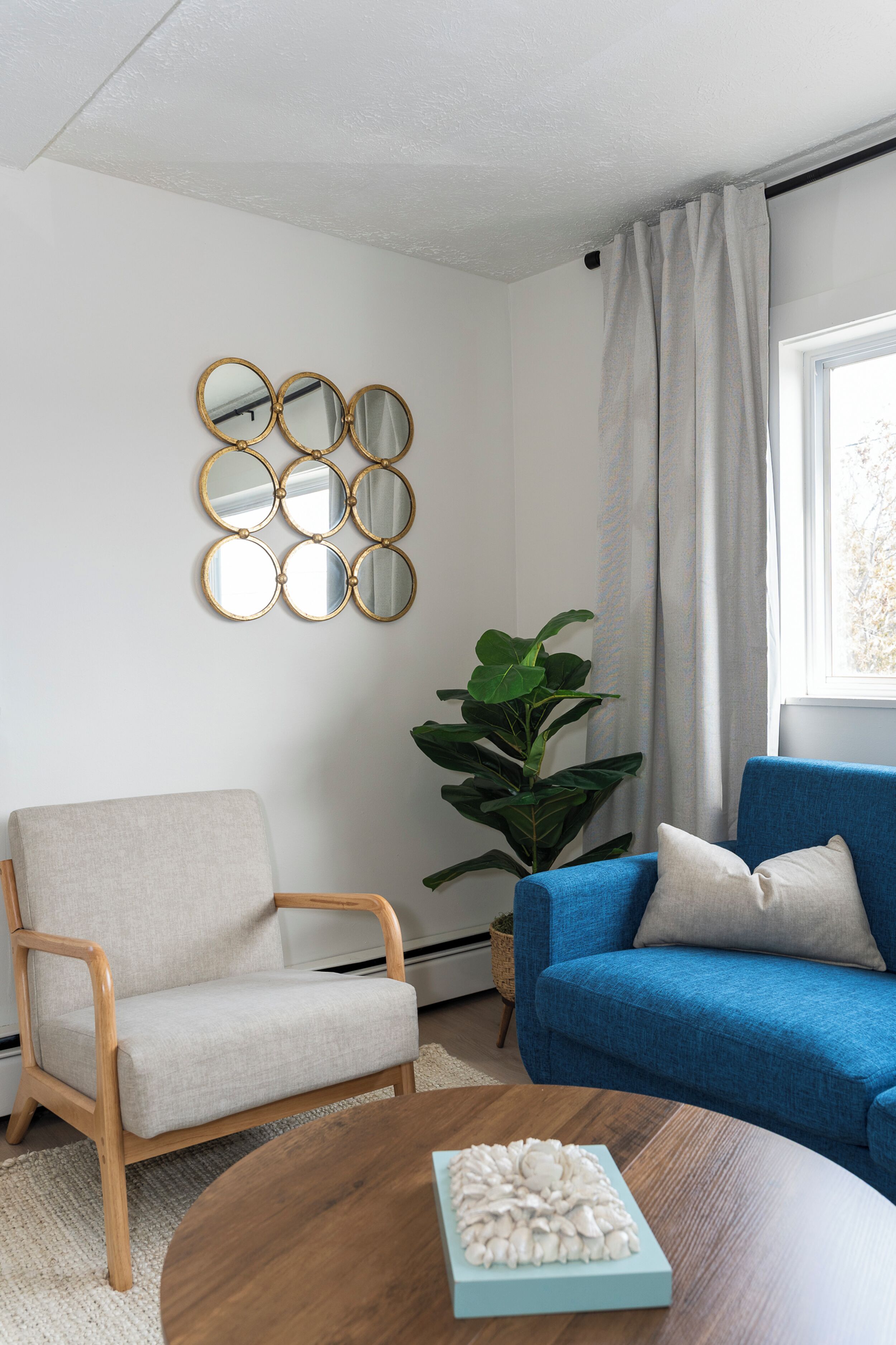 Un salon avec un fauteuil blanc, un canapé bleu, une table basse ronde ornée d’un objet décoratif et une grande plante verte.