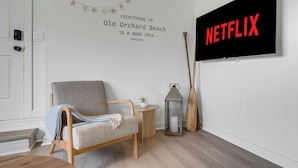 A living room with a grey sofa, a small side table with a plant, a wooden floor, and a wall-mounted TV displaying 'NETFLIX'.