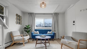 A modern living room with a blue sofa, two wooden chairs, a round coffee table, and a large window with curtains.