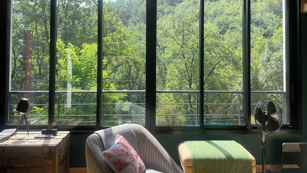 Una habitación con un ventanal grande, una silla, un escritorio con una computadora portátil, un ventilador y un cojín verde.