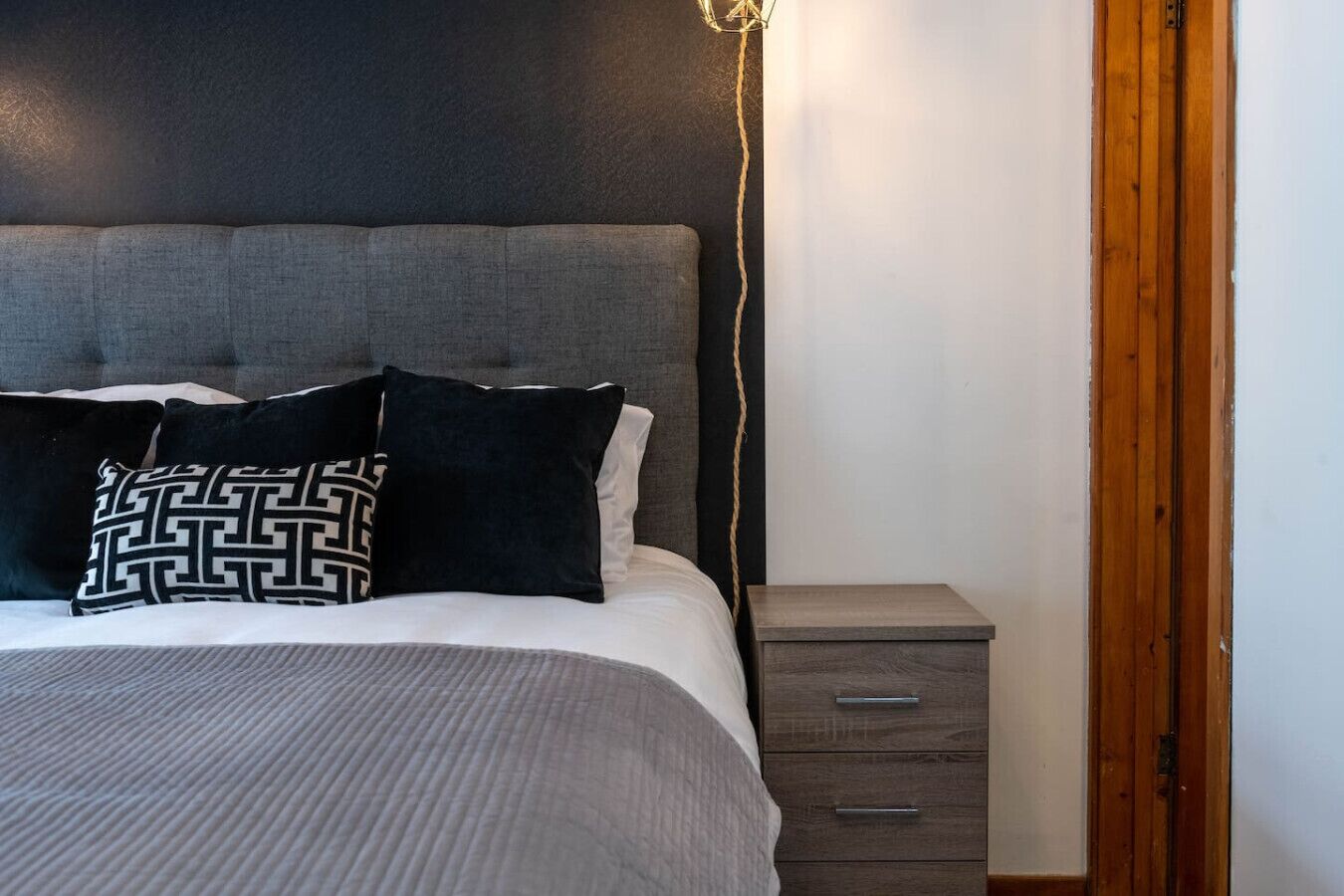 Una cama con cabecera gris, una mesita de noche con cajones y una lámpara de pared.