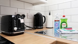 Et køkken med kaffemaskine, elkedel, en rulle køkkenrulle, opvaskesæbe og en rengøringsklud på bordet.
