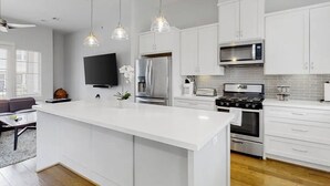A modern kitchen with white cabinetry, a central island, stainless steel appliances, pendant lighting, and a flat-screen TV. - Urban Escape in the heart of Downtown Houston (Houston)