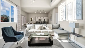 Ein modernes Wohnzimmer mit einer Couch, einem Sessel und einem Couchtisch, im Hintergrund eine Küche und große Fenster.
