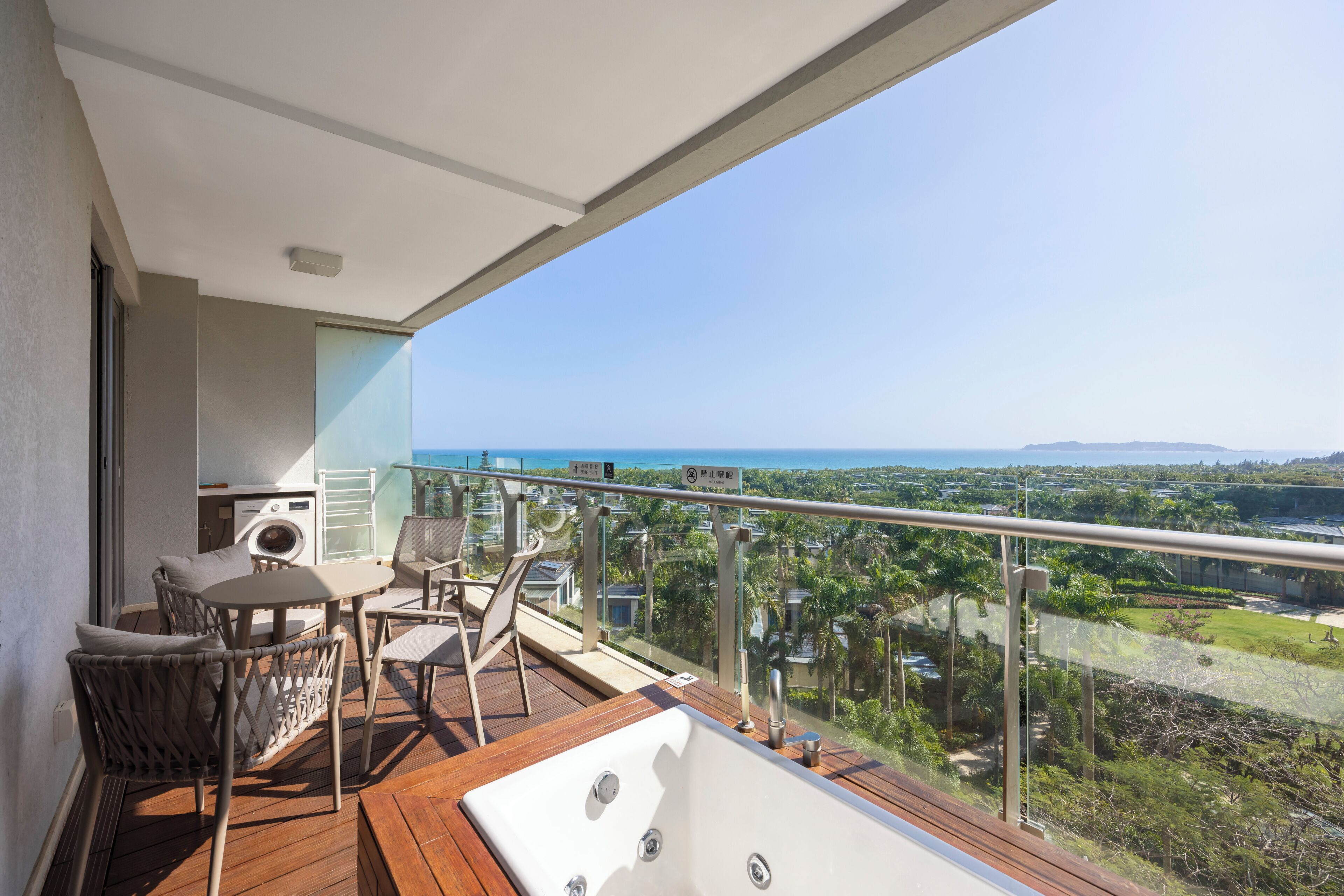 Een balkon met een houten terras, een zithoek met stoelen en een tafel, en uitzicht op zee en groen.