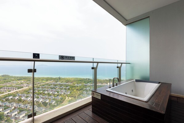 Een balkon met uitzicht op een woonwijk en de zee, een badkuip en een glazen hekwerk.