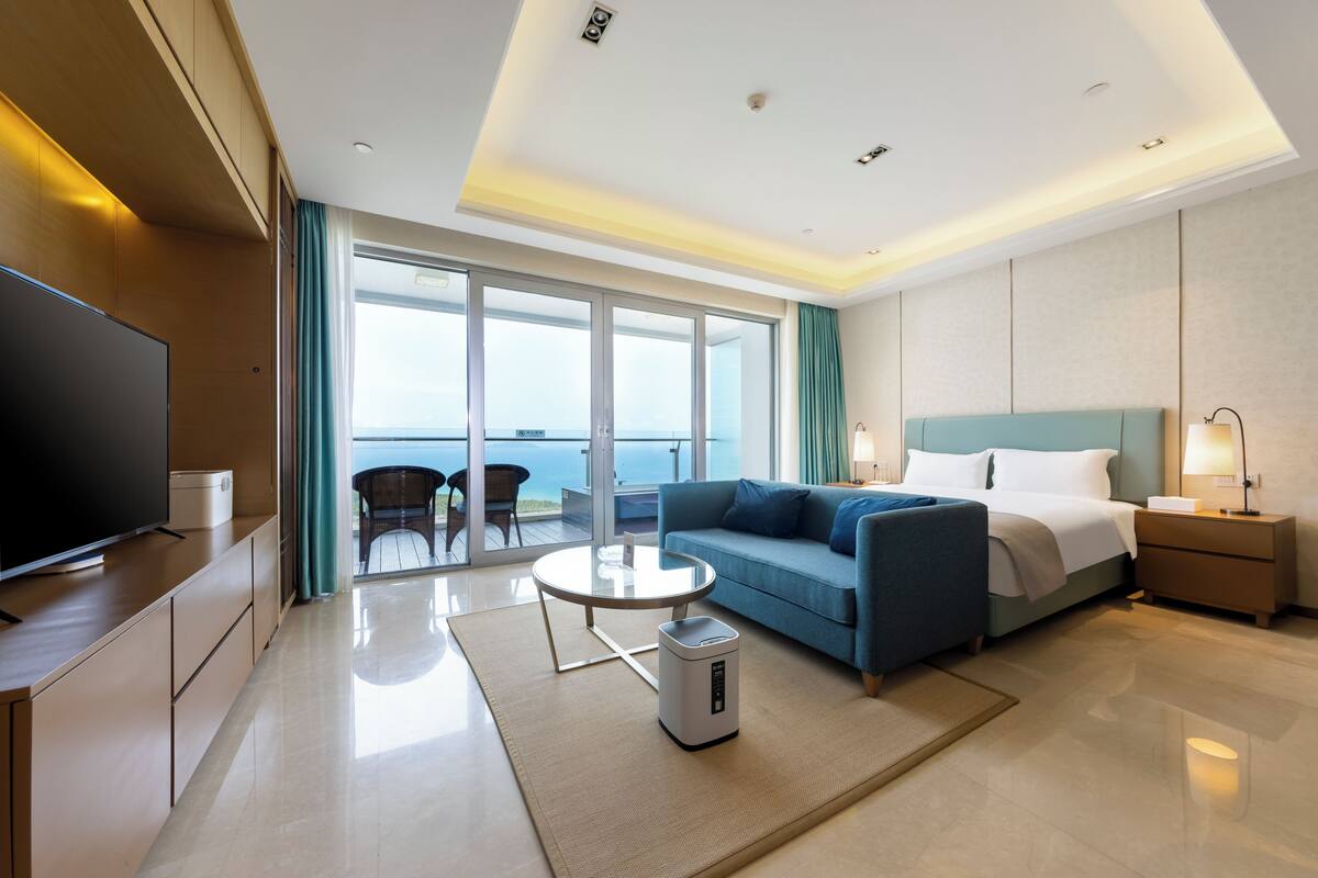 Una habitación de hotel moderna con un televisor grande, un sofá, una cama, una mesita de noche, una lámpara y vistas al océano a través de una puerta corrediza de vidrio.