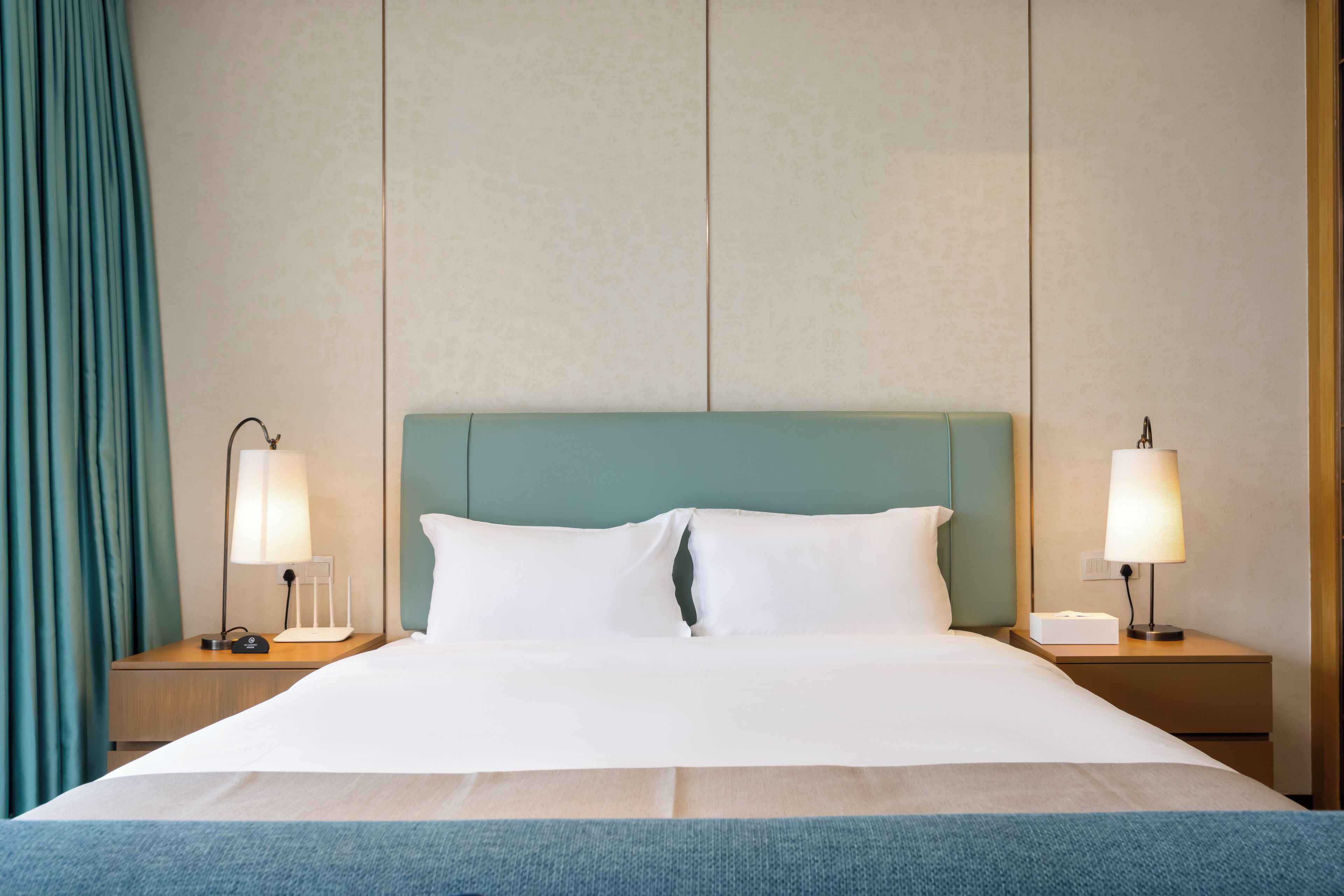 Hotellihuone, jossa on suuri sänky, kaksi yöpöytää valaisimineen, turkoosinsininen pääty ja verho vasemmalla puolella.