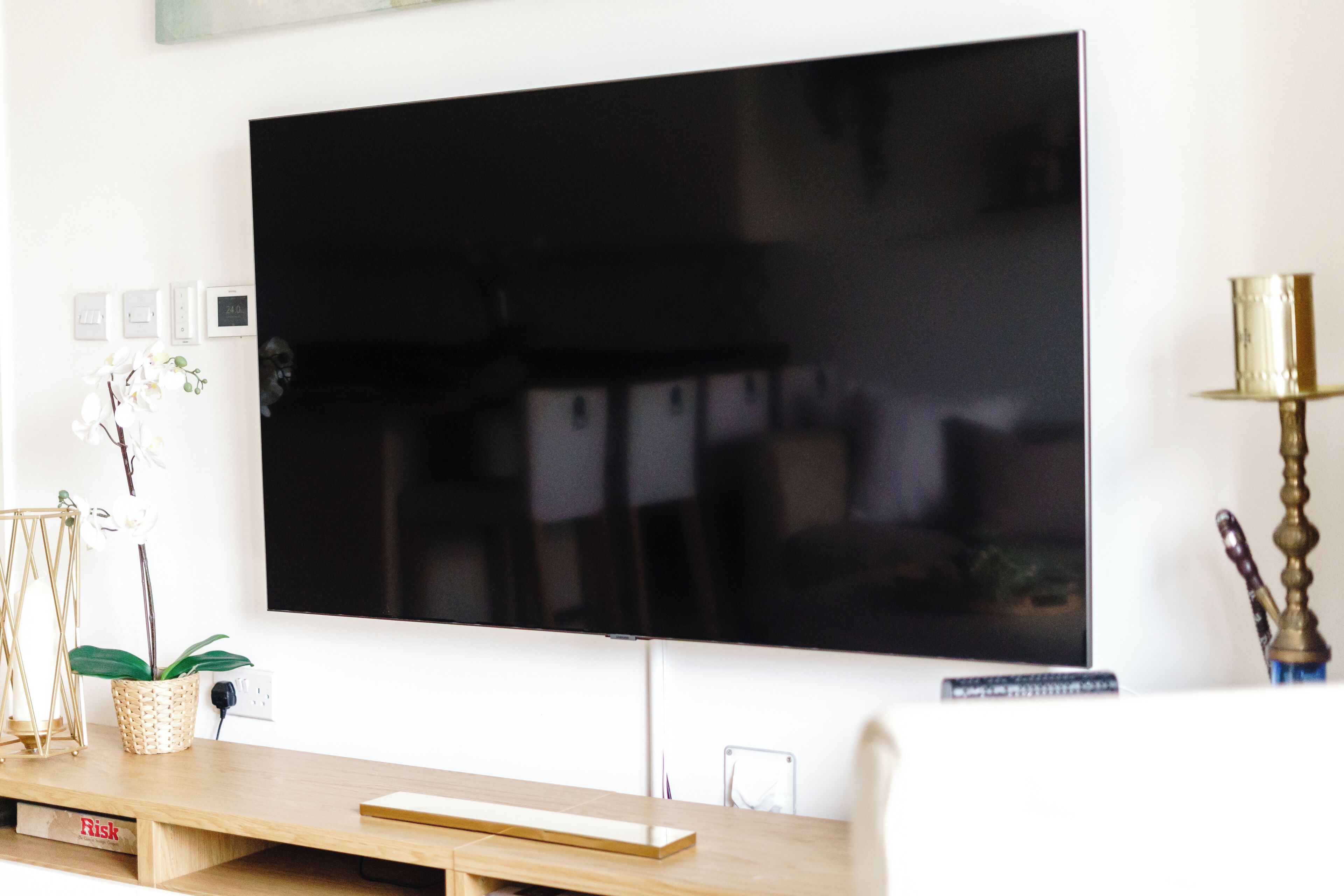 A flat-screen TV mounted on a white wall, a wooden side table with a remote control, a small plant, and a decorative lamp.