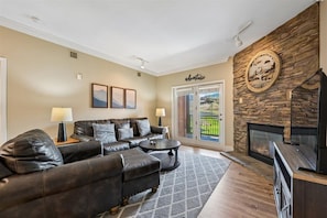 A living room with a stone fireplace, a leather sofa, a coffee table, and a view through a glass door to a balcony. - Baskins Creek 414 🌲 Walk to Downtown Gatlinburg | Pool, Hot Tub + Fireplace 🔥 (Gatlinburg)