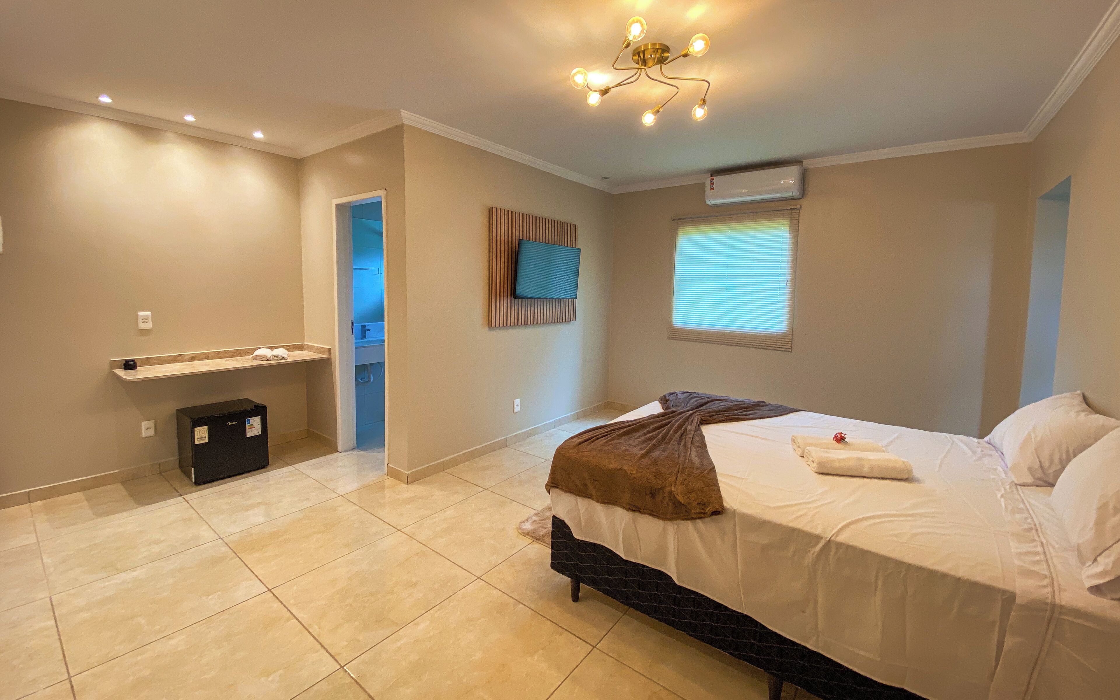 Un dormitorio con una cama, un televisor montado en la pared, una ventana con persianas y una lámpara en el techo.