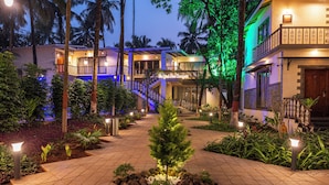 A well-lit courtyard with a central tree, surrounded by plants and a tiled walkway leading to a building with multiple balconies. - Parkwood Exotica Resort (Alibag)