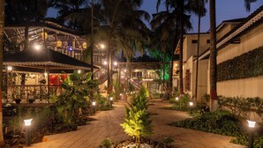 A well-lit outdoor area with a pathway, palm trees, and a building with a balcony. - Parkwood Exotica Resort (Alibag)