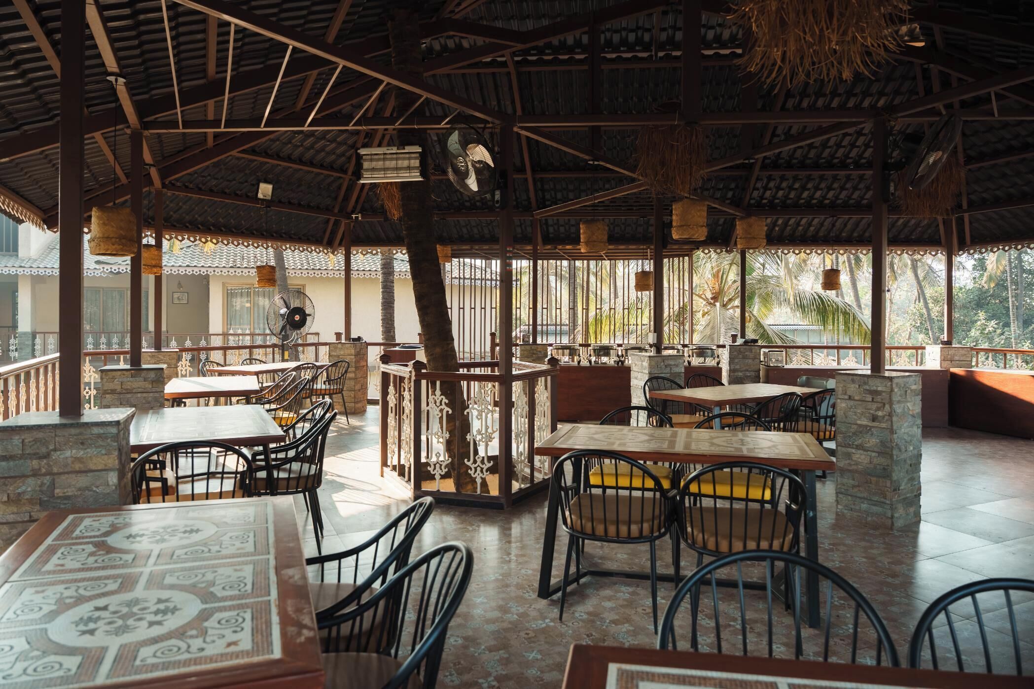 Restaurante ao ar livre com mesas e cadeiras, telhado de palha e vista para palmeiras.
