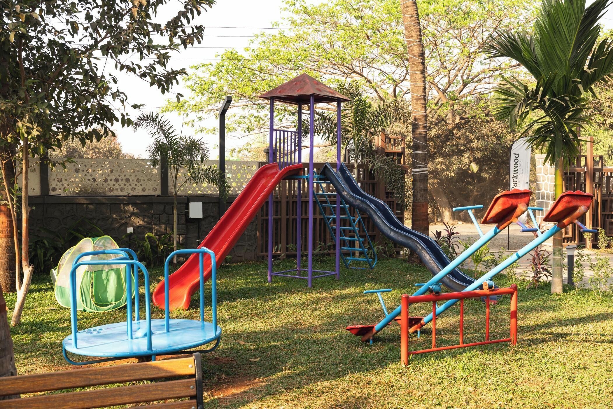A playground with slides and swings, surrounded by trees and a stone wall.