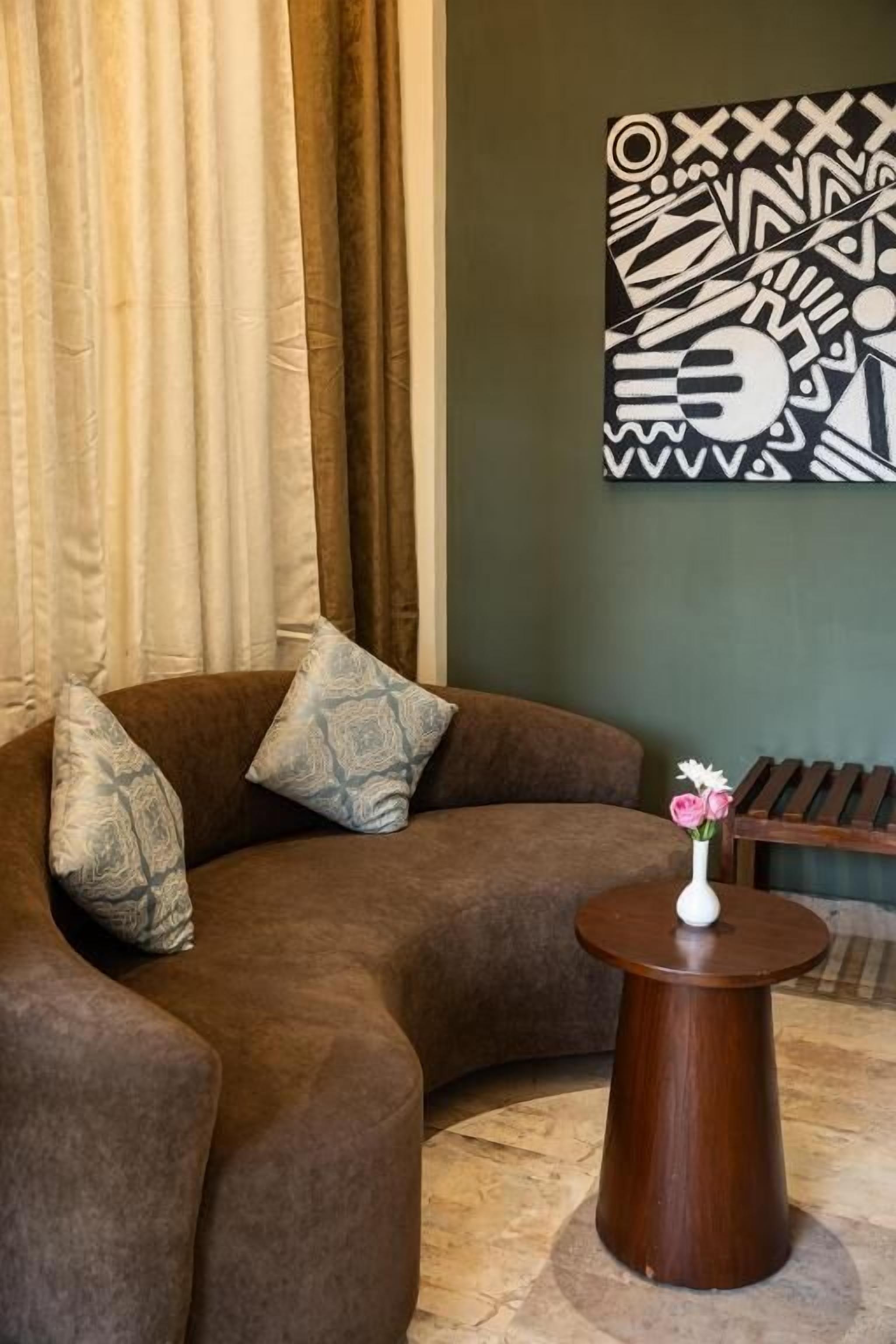 Un sofá marrón con cojines estampados, una mesita redonda de madera con un jarrón de flores y una obra de arte geométrica en la pared.