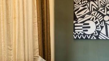Een bruine bank met gekleurde kussens, een rond houten bijzettafeltje met een vaas bloemen, en een wanddecoratie met een geometrisch patroon.