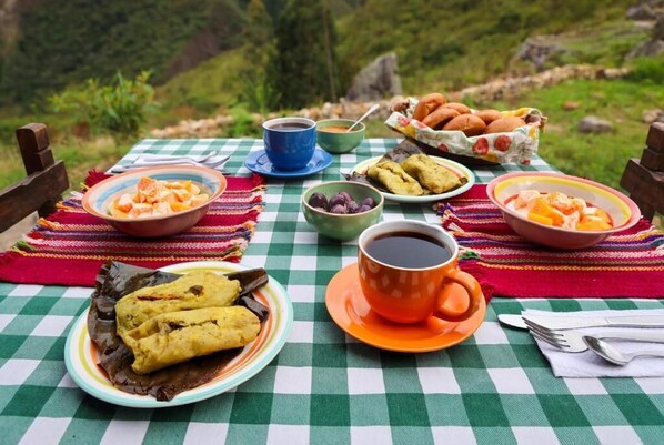 Une table dressée à l’extérieur, avec différents plats, des tasses et une nappe à carreaux.