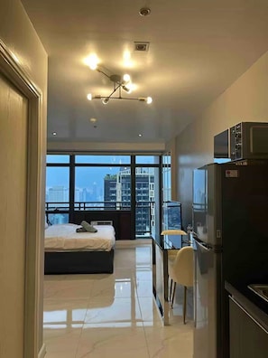 Une chambre d’hôtel moderne équipée d’un lit, d’un bureau avec une chaise, d’un réfrigérateur et offrant une vue sur le paysage urbain grâce à de grandes fenêtres.